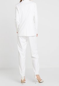 Dopasowana biała marynarka z pojedynczym rozcięciem, w połączeniu z dopasowanymi spodniami. Charakteryzuje się gładką tkaniną i elegancką sylwetką, dopełnioną beżowymi szpilkami.