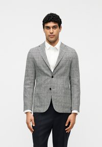 Άνδρας που φοράει γκρι καρό σακάκι με δύο κουμπιά πάνω από λευκό πουκάμισο και μαύρο παντελόνι, στέκεται μπροστά σε απλό λευκό φόντο.