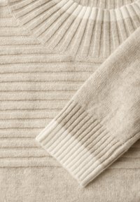 Beiger Strickpullover mit geripptem Muster an der Passe und den Ärmeln, mit kontrastierenden, gerippten Bündchen. Weicher Stoff mit leichter Glanz.