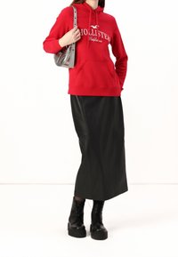 Sudadera roja con el texto "Hollister California", combinada con una falda larga de cuero negra. Completado con botas de plataforma negras y un bolso plateado.