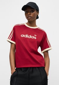 Νέα γυναίκα που φοράει κόκκινο δίχτυ Adidas με εκρού περιθώριο και μαύρο καπέλο, στέκεται με τα χέρια στις τσέπες μαύρου παντελονιού.