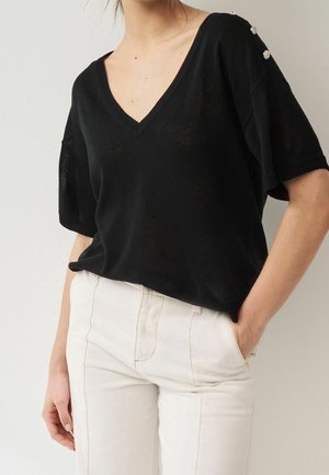 Vrouw draagt een zwarte V-hals korte mouwen trui met decoratieve knopen op de schouder en witte broek, hand in de zak.