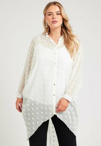 Blusa blanca transparente con patrón de lunares texturizados, cierre frontal con botones, cuello y diseño de dobladillo asimétrico; mangas largas y tela ligera.