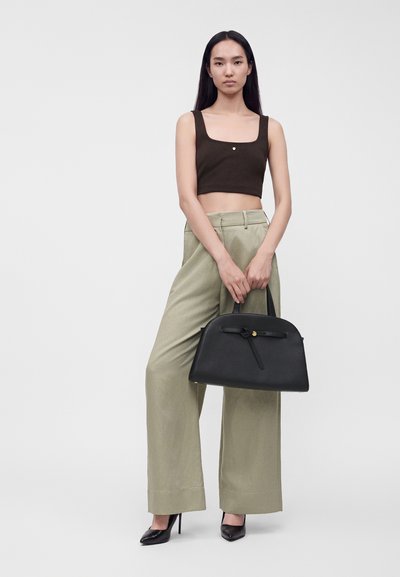 Γυναίκα με μαύρο crop top και μπεζ φαρδιά παντελόνια κρατώντας μια μεγάλη μαύρη τσάντα, στέκεται μπροστά σε απλό φωτεινό φόντο.