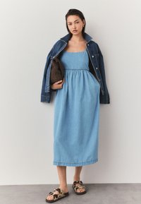 Femeie purtând o rochie midi din denim albastru deschis cu buzunare, o jachetă din denim închis pe umeri, ținând o geantă tricotată maro, purtând sandale cu model.