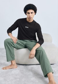 Cămașă neagră cu mâneci lungi și logo-ul "BOSS" alb, asortată cu pantaloni verzi cu carouri, stând pe un puf rotund și moale, de culoare crem.