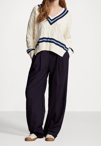 Krämfärgad stickad tröja med blå och marinblå ränder, V-hals och kabelmönster. Matchad med löst sittande marinblå byxor och vita sneakers.