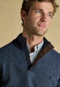 Tumši zils vilnas džemperis ar teksturētu audumu, adītu apkakli un brūnu ādas akcentu pie rāvējslēdzēja. Zem tā valkāts svītrains krekls.