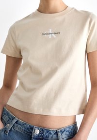 Persona che indossa una maglietta corta beige con il logo "Calvin Klein Jeans" e jeans di denim blu, mostrando la pancia e con le mani in tasca.