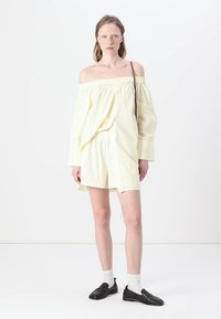 Off-shoulder lys gul topp og matchende shorts i myk stoff. Modellen har på seg svarte slip-on sko og hvite sokker, sammen med en enkel skulderveske.