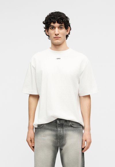 Άνδρας με σγουρά σκούρα μαλλιά, που φοράει άνετη λευκή μπλούζα με το κείμενο "OFF" και ξεθωριασμένο γκρι τζιν, στέκεται μπροστά σε απλό φωτεινό φόντο.