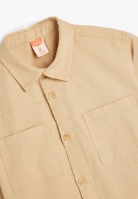 Camicia beige a maniche corte in cotone con colletto, due tasche sul petto e quattro bottoni, caratterizzata da una texture liscia e cuciture uniformi.