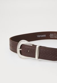 Curea din piele maro cu textură împletită, cataramă din metal argintiu și mai multe găuri de ajustare. Eticheta spune "Vanzetti, Fabricat în Germania."
