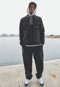 Giacca funzionale nera con grandi tasche, indossata sopra un hoodie grigio. Pantaloni neri larghi e sneakers bianche con accenti neri e rossi.
