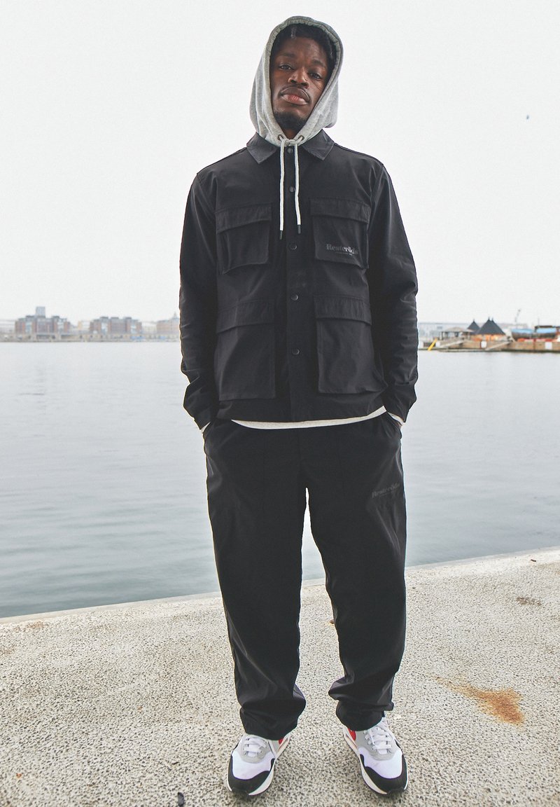Giacca funzionale nera con grandi tasche, indossata sopra un hoodie grigio. Pantaloni neri larghi e sneakers bianche con accenti neri e rossi.