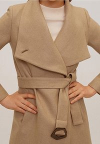 Manteau beige croisé avec un large col et une ceinture à nouer, orné d'une boucle accentuée foncée. Confectionné dans un tissu texturé.