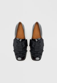 Chaussures en cuir verni noir au design plat, ornées d'un grand nœud et d'accents de rubans superposés à l'avant. Semelle intérieure marron texturée visible.