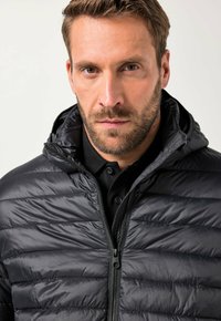 Sort pufferjakke med vandret quiltning, stående krave og lynlåslukning. Bæret over en sort polo shirt, har en elegant, glat tekstur.