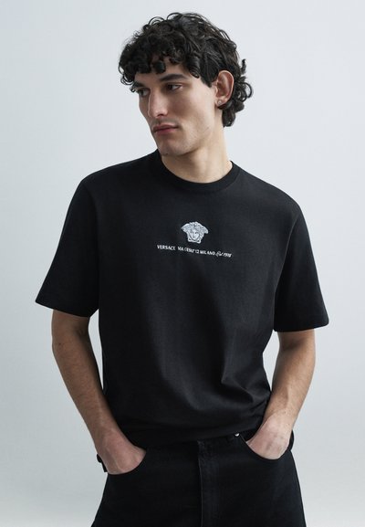 T-shirt en coton noir avec un motif blanc centré représentant une tête de Méduse et du texte, doté d'un col rond et de manches courtes.