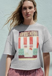 T-shirt grigio corto con un design grafico colorato che rappresenta un negozio. Strisce rosse e bianche, accenti verdi, maniche corte e vestibilità rilassata.