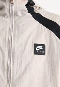 Nike jacket in light beige fabric with a full zip closure, black mesh accents, and a square logo patch displaying "NIKE AIR."