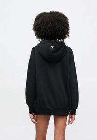 Göndör hajú személy, aki fekete, túlméretezett kapucnis pulóvert visel, háttal áll, egyszínű fehér háttér előtt.