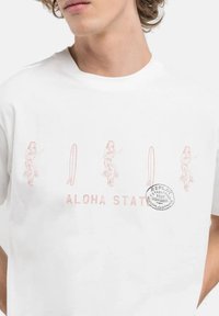 Witte katoenen T-shirt met roze graphics van hula dansers en een surfplank, met de tekst "ALOHA STATE" en een ronde logo-accent.