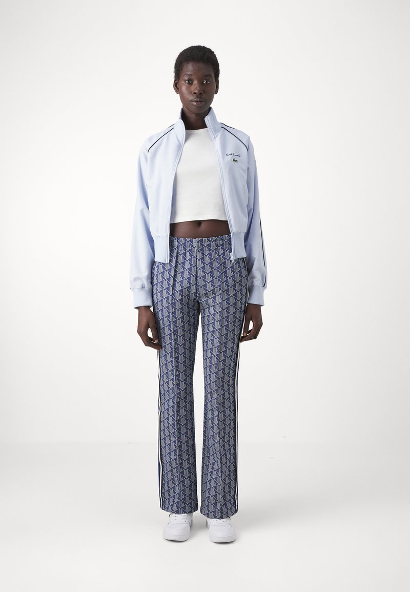 Jasnoniebieska kurtka zapinana na zamek z czarnymi akcentami, połączona z wzorzystymi niebieskimi spodniami w pionowe paski. Modelka nosi białą krótką bluzkę oraz trampki.