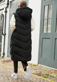 Czarna pikowana kamizelka z kapturem, z wzorem w jodełkę. Połączona z dopasowanymi czarnymi spodniami i białymi trampkami, spacerując po brukowanym chodniku.