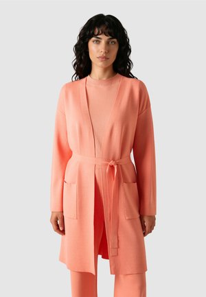 Frau mit dunklen lockigen Haaren, die eine pfirsichfarbene Strickjacke trägt, die in der Taille gebunden ist, über einem passenden pfirsichfarbenen Oberteil und Hose, stehend vor einem einfarbigen Hintergrund.