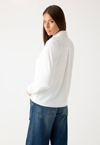 Weiße Bluse mit langen �Ärmeln und strukturiertem Kragen. Kombiniert mit blauen, weiten Denim-Jeans. Glatter Stoff, lässige Passform und Knopfdetails.