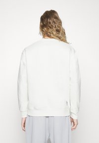 Sweat-shirt blanc à manches longues et col rond, en tissu texturé avec un petit détail logo sur le côté inférieur.