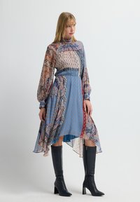 Mujer con un vestido midi azul de estampado, de cuello alto y mangas largas, con un dobladillo asimétrico y botas negras de tacón alto hasta la rodilla, de pie.