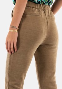 Pantaloni in corduroy marroni con righe verticali, vita alta e due tasche posteriori, mostrati di lato con una mano appoggiata sui fianchi.