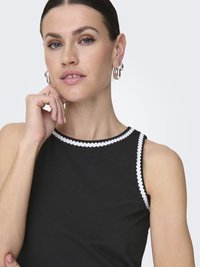 Čierny bezrukávový top so zúbkovaným bielym lemom okolo výstrihu. Modelka má krátke vlasy a nosí strieborné kruhové náušnice. Pleť je svetlá.