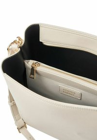 Sac à main synthétique beige avec une fermeture éclair dorée, un intérieur noir et une poche avant étiquetée "ÉNERGIE POSITIVE". Bandoulière ajustable incluse.