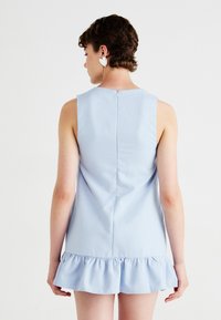 Light blue sleeveless dress with a ruffled hem, featuring a back zipper closure and a smooth texture. Earphones visible on model.