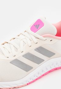 Beige athletic shoe featuring a mesh upper, textured grey stripes, and a pink accent at the heel. Laces are white and the sole has a pink detail.