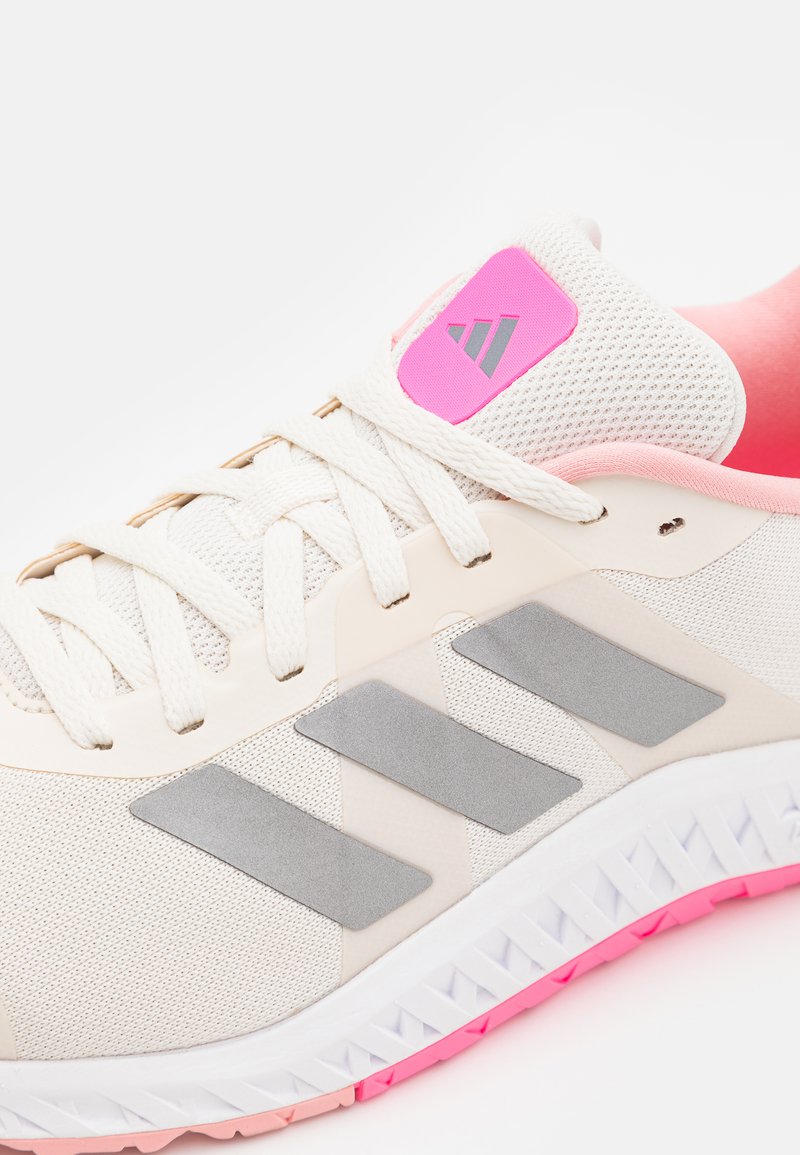 Zapatilla deportiva beige con parte superior de malla, franjas grises texturizadas y un detalle rosa en el talón. Los cordones son blancos y la suela tiene un detalle rosa.