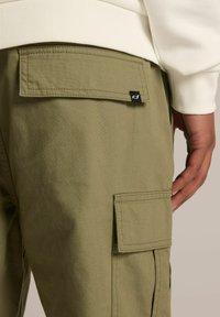 Oliwkowe bawełniane spodnie cargo mają tylną kieszeń naszywaną z małą czarną metką, podkreślającą mocny materiał i praktyczny design.