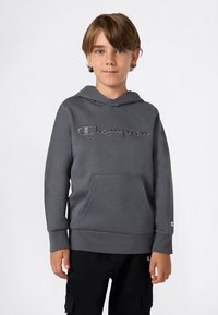 Ragazzo con capelli castano chiaro che indossa un felpa grigia Champion e pantaloni cargo neri, in piedi su uno sfondo bianco uniforme.