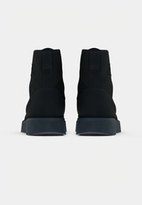 Botas negras hasta el tobillo con material suave, puntera redondeada y suela oscura resistente. Presenta un tirador en la parte posterior y un detalle de costura sutil.