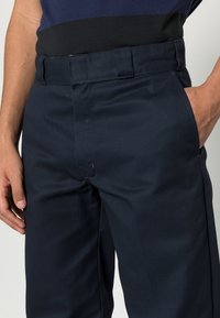Homme portant un pantalon bleu marine foncé et une chemise noire et bleue foncée rentrée, avec une main dans la poche et l'autre reposant sur le côté.