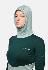 Frau in einem grünen und hellgrauen, mit Vaude-Logo versehenen, thermischen Kapuzen-Sportoberteil, die nach links schaut und einen neutralen Gesichtsausdruck hat, vor einem weißen Hintergrund.