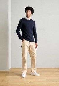 Navy sweater with a round neckline over a white collared shirt, paired with beige cargo pants and white sneakers, set on a wooden floor.
