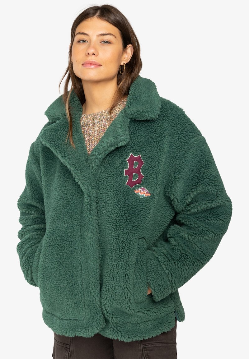 Grön fleecejacka med en plyschliknande textur, oversized design och skuren krage. Har en vinröd broderad logotyp på vänster sida.