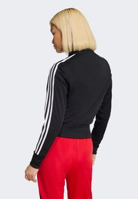 Fekete atlétikai dzseki fehér csíkokkal az ujjain, testhez simuló derékrésszel és sima textúrával; piros atlétikai nadrággal párosítva.