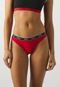Czerwone majtki bikini z czarnym pasem, na którym widnieje białe napisy "HUGO". Gładki materiał, niski krój i minimalne zakrycie.
