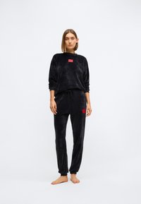 Conjunto de roupa de lounge em veludo preto, apresentando uma t-shirt de gola redonda com um etiqueta vermelha e calças ajustadas a combinar com bolsos laterais e punhos elásticos.