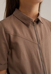 Camisa corta de manga corta de algodón marrón con cierre de cremallera, con un collar puntiagudo, tejido texturizado y una costura diagonal en el pecho.